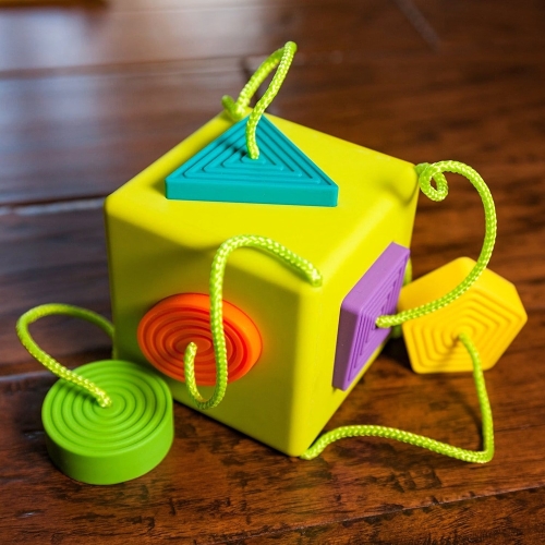 Sorter Kostka OombeeCube Fat Brain Toys na drewnianym stole – zielona kostka z kolorowymi kształtami trójkąt, koło i kwadrat na sznurkach