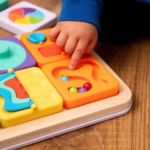 Tablica PlayTab Fat Brain Toys – zbliżenie na dłoń niemowlęcia dotykającą pomarańczowego panelu sensorycznego z kolorowym labiryntem z kuleczkami, zabawka rozwijająca motorykę małą dla dzieci od 6 miesięcy