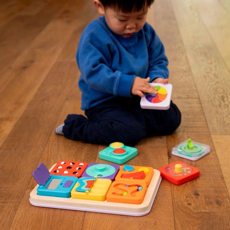 Tablica PlayTab Fat Brain Toys – maluch siedzący na podłodze ogląda biały panel z kolorowym, obrotowym kołem tęczowym, obok rozłożona tablica z pozostałymi modułami, zabawka sensoryczna rozwijająca kreatywność