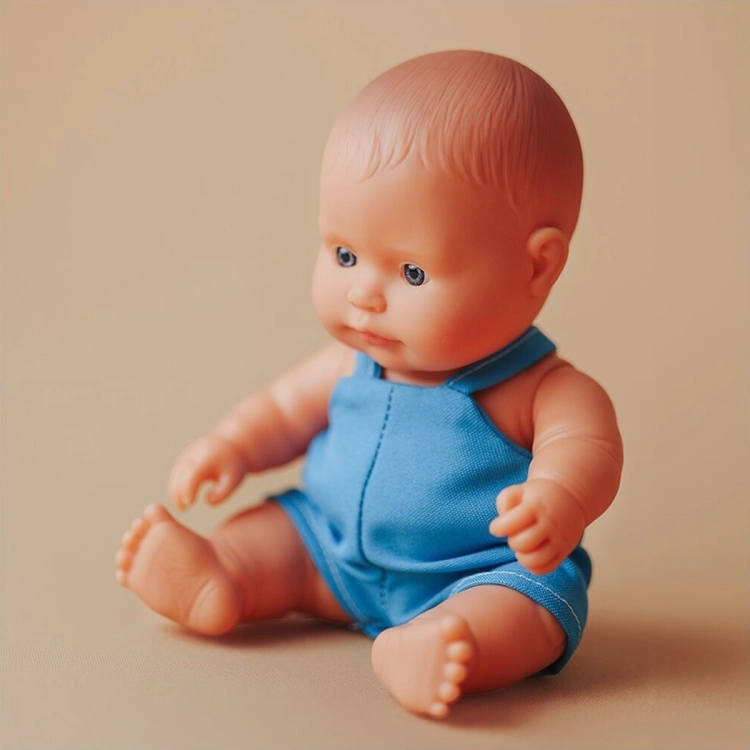 Lalka Miniland Baby chłopiec 21 cm w niebieskich ogrodniczkach, idealna do małych rąk, na ciepłym beżowym tle