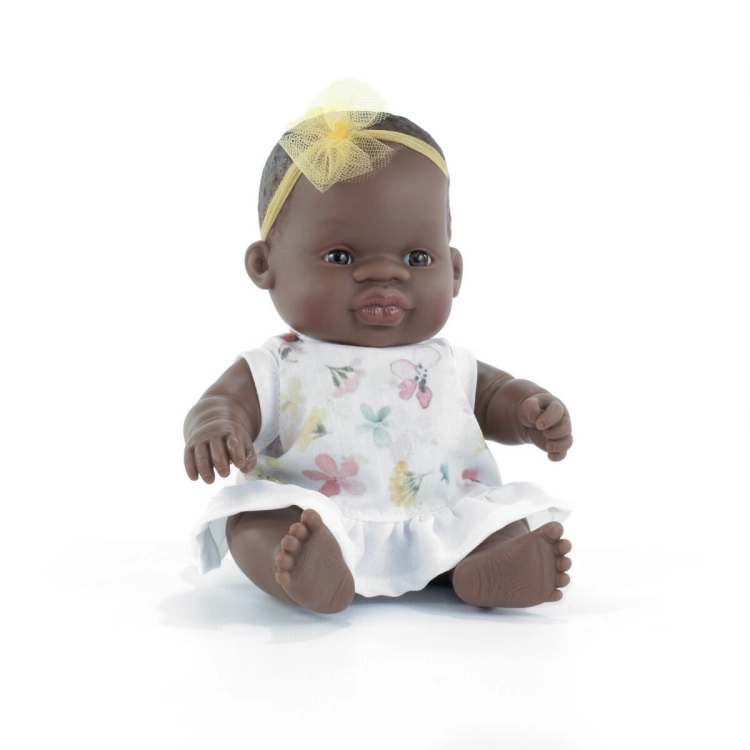 Lalka bobas dziewczynka Miniland Baby 21 cm o ciemnej karnacji, w białej sukience w kwiaty i żółtej opasce, na białym tle