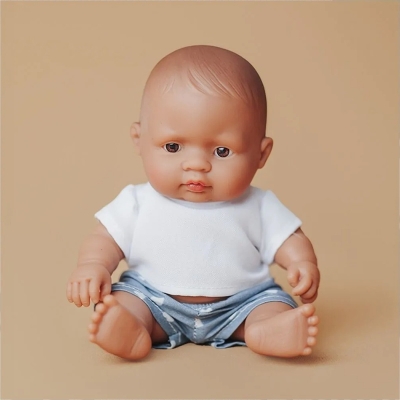 Mała lalka chłopiec Miniland 21 cm o brązowych oczach, w białym T-shircie i niebieskich szortach, siedząca na beżowym tle