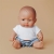 Mała lalka chłopiec Miniland 21 cm o brązowych oczach, w białym T-shircie i niebieskich szortach, siedząca na beżowym tle