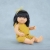 Lalka Miniland 38 cm o azjatyckich rysach, w musztardowym kombinezonie i żółtej opasce z tiulu, siedząca na błękitnym tle